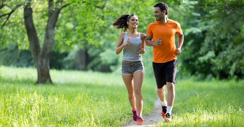 Un homme et une femme courent (font du sport) ensemble dans un parc verdoyant, souriant et motivé.